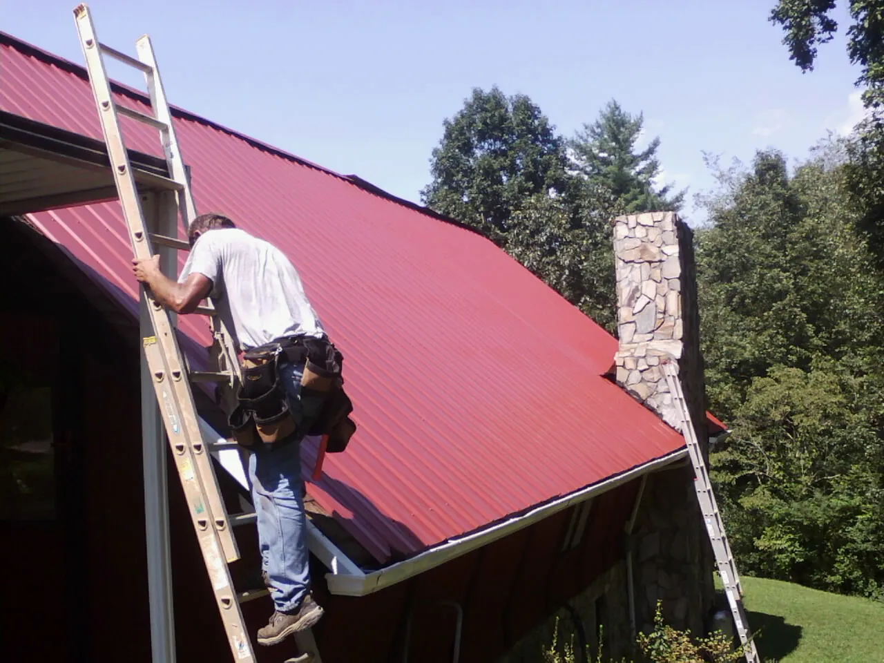 Roofer on a ladder beside a red metal roof and stone chimney — Soffit & Fascia Repair by Roofing Brothers in Laverne